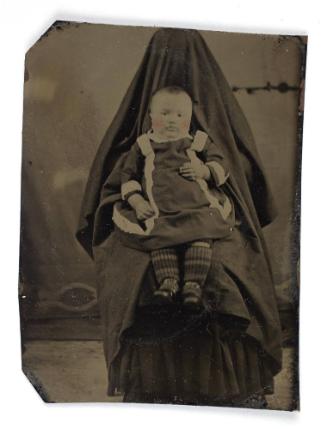 Portrait of baby wearing jacket with white trim (seated on lap of shrouded figure)