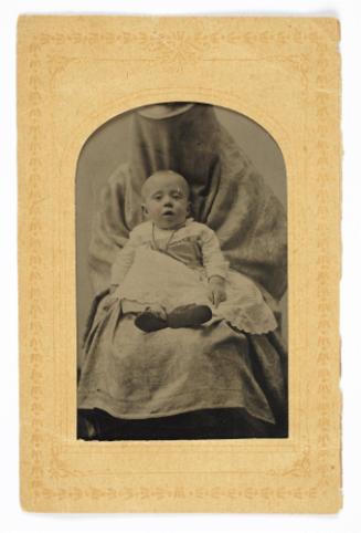 Portrait of baby wearing necklace (seated upon lap of shrouded figure)