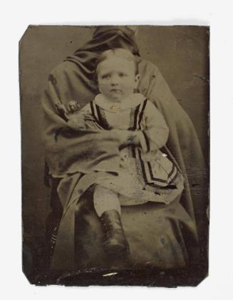 Portrait of a young girl wearing boots (seated on lap of shrouded figure)