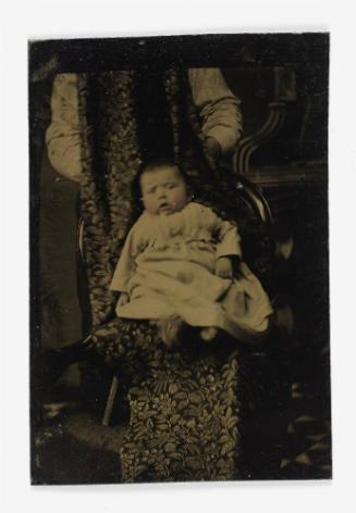 Portrait of a baby (seated on chair, supported by standing figure)