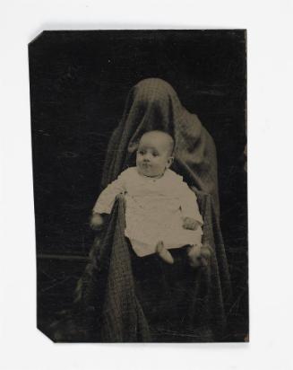 Portrait of baby in white (seated on lap of shrouded figure)