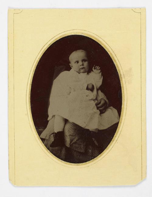 Portrait of baby wearing white dress and boots ( supported on lap of figure)