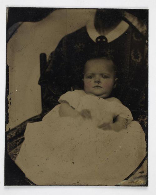 Portrait of baby in white dress (seated on lap with cropped face of woman)