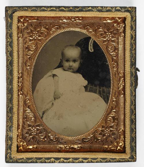 Portrait of baby in white dress (seated on lap of cropped figure)